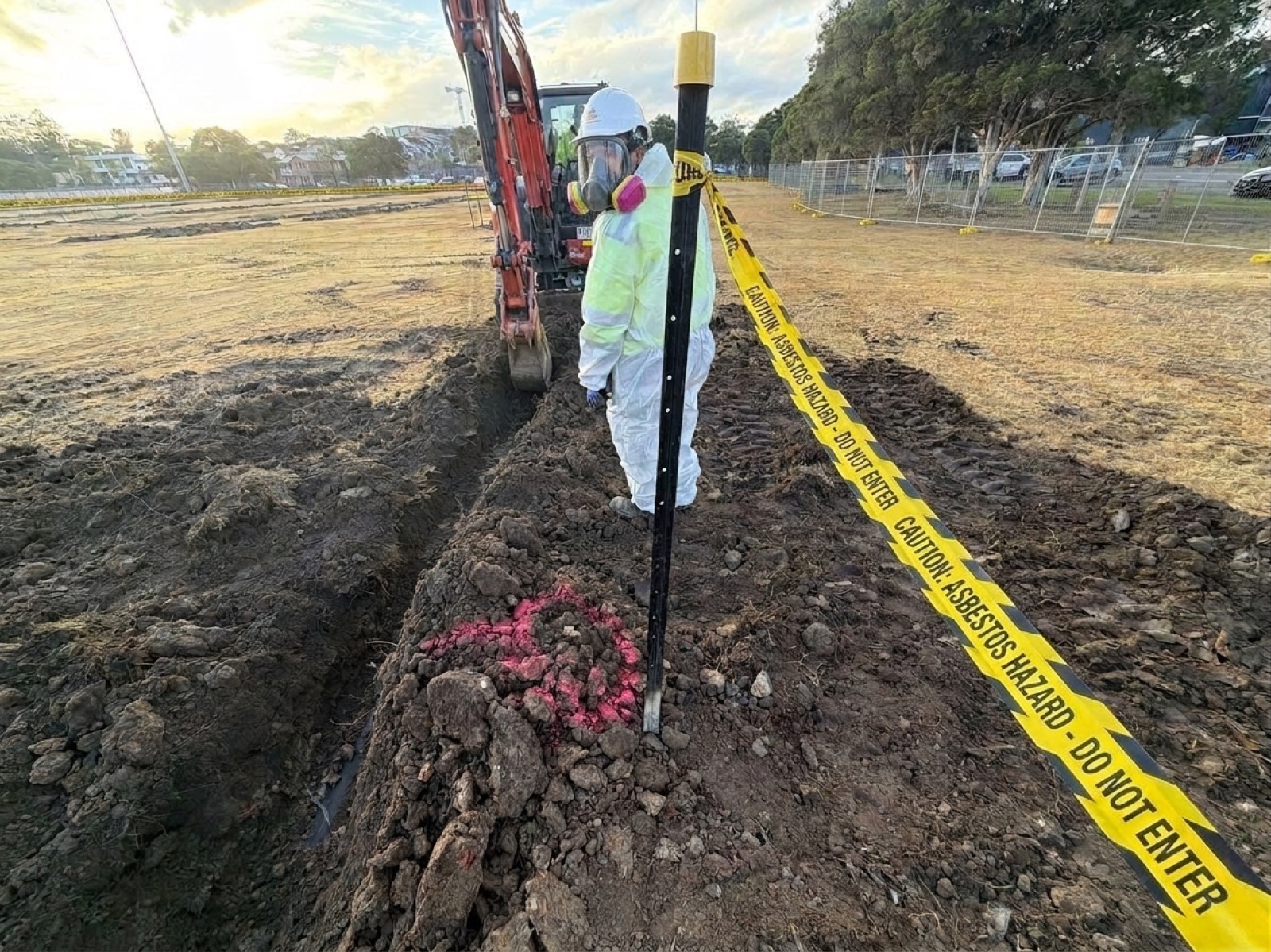 Suited operator beside excavation trench behind asbestos hazard tape, Doyle Ground Parramatta